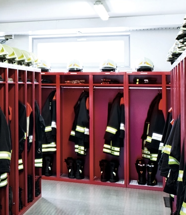 Feuerwehrjacken und Helme in roten Spinden in einem Umkleideraum.